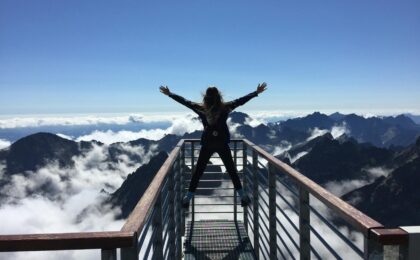 person standing on hand rails with arms wide open facing the mountains and clouds