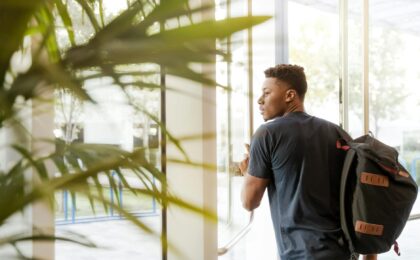 man looking outside window carrying black and brown backpack while holding his hand on window